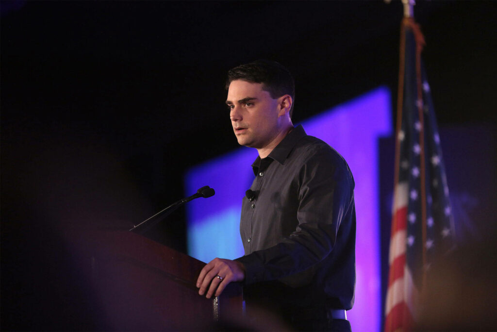 Ben Shapiro speaking at podium with American flag and dramatic lighting wallpaper background