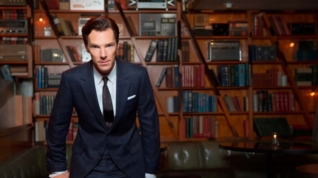 Benedict Cumberbatch in stylish suit stands confidently before bookshelves wallpaper background