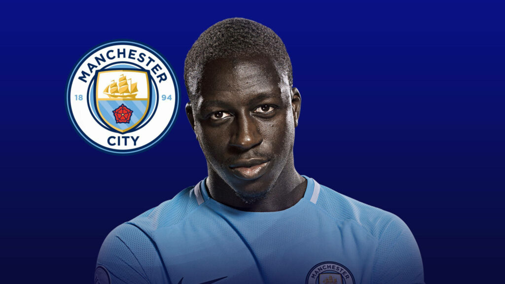 Benjamin Mendy in Manchester City kit standing before club logo on deep blue gradient wallpaper