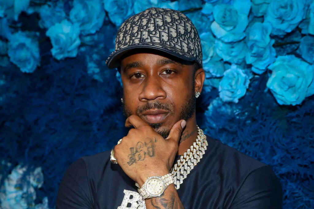 Benny The Butcher confident with flashy jewelry against vibrant blue rose wallpaper background