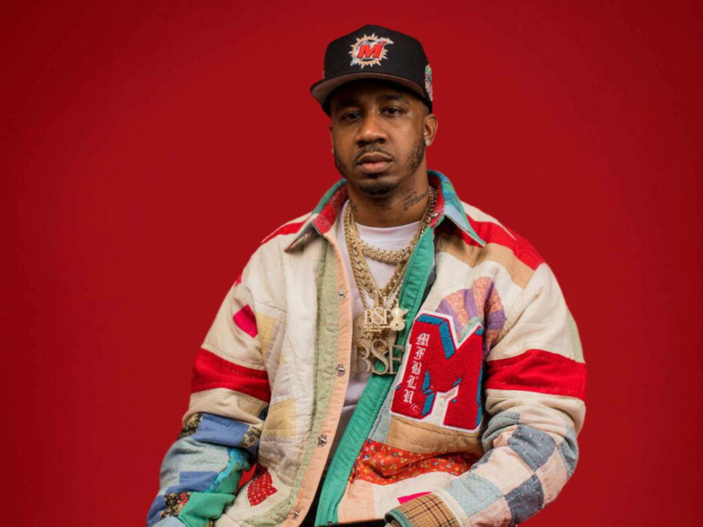 Benny The Butcher portrait wallpaper rapper in colorful jacket gold chains black cap red background