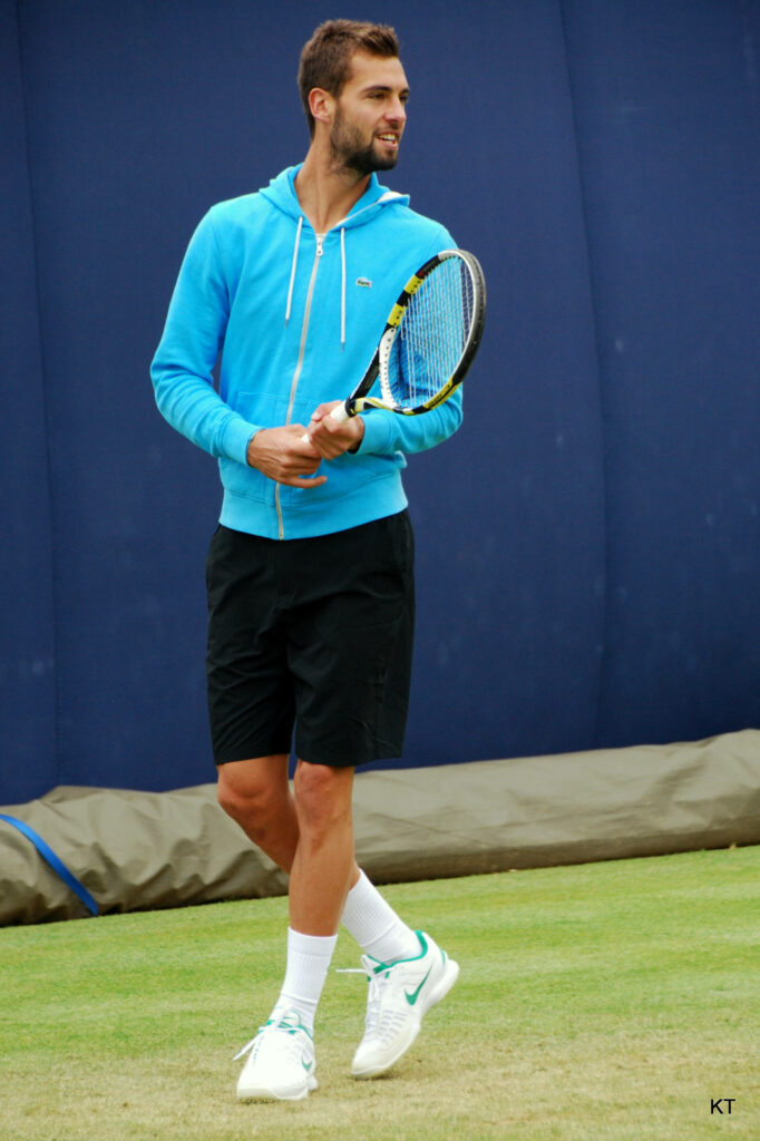 Benoît Paire on grass tennis court in blue hoodie holding racket legs crossed against wallpaper background