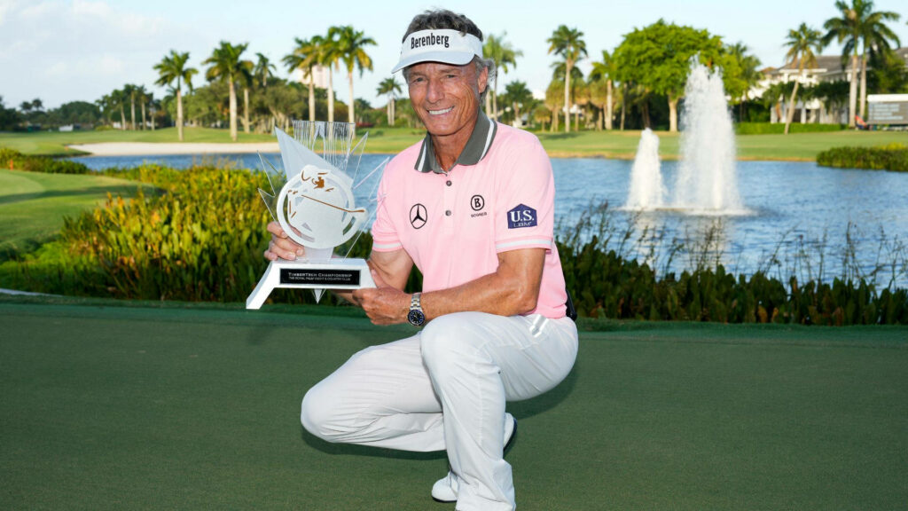 Bernhard Langer crouching on golf course near lake holding trophy smiling with palm trees fountain Wallpaper