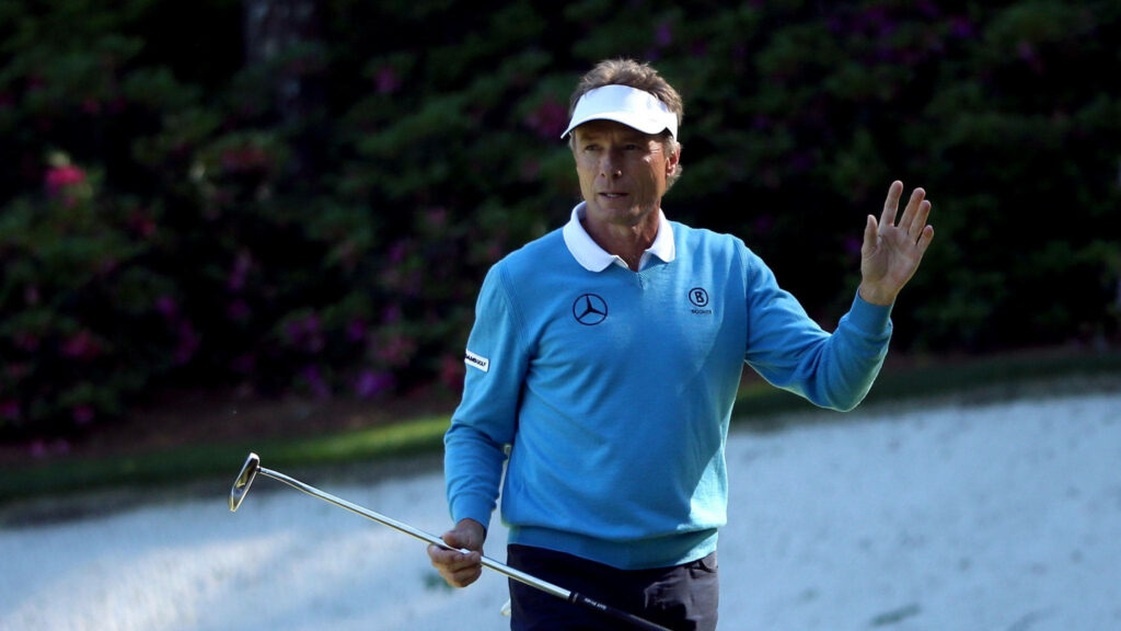 Bernhard Langer waving on golf course in blue sweater visor holding putter calm expression wallpaper