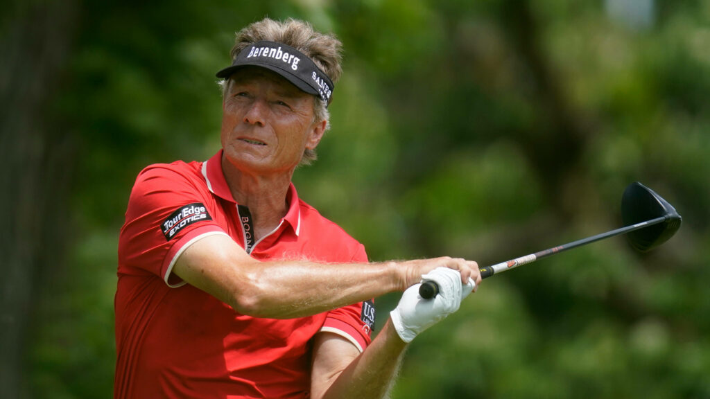 Bernhard Langer mid-swing on golf course wearing red shirt visor with green foliage background Wallpaper