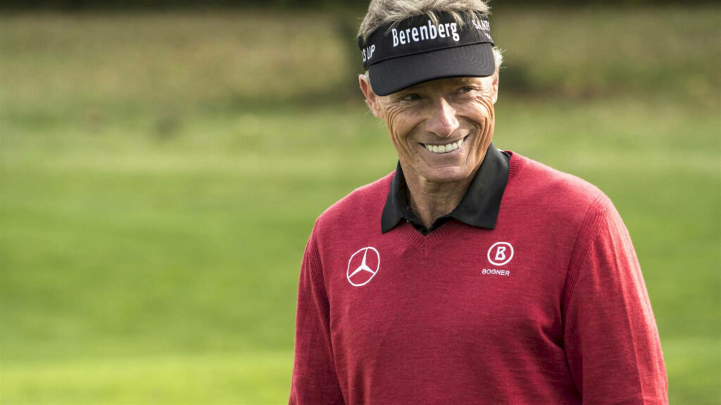 Bernhard Langer smiling on golf course wearing red sweater and signature visor with green background Wallpaper