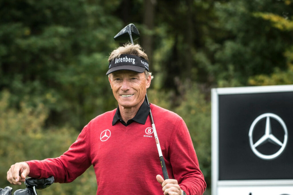 Bernhard Langer smiling on golf course wearing red sweater and black visor with Mercedes-Benz sign Wallpaper