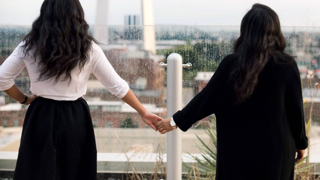 Two friends with long dark hair holding hands on rainy rooftop with Washington Monument view Wallpaper