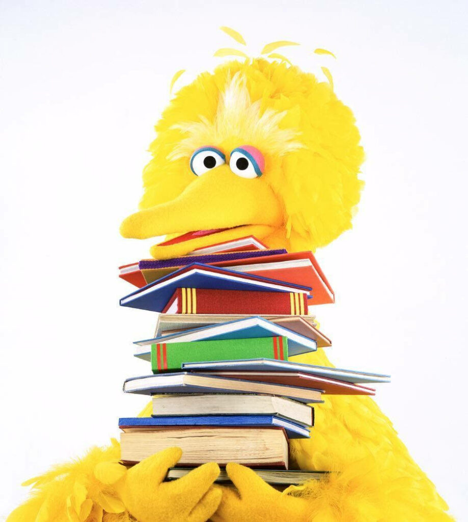 Big Bird cheerfully holding tall colorful book stack vibrant playful wallpaper background