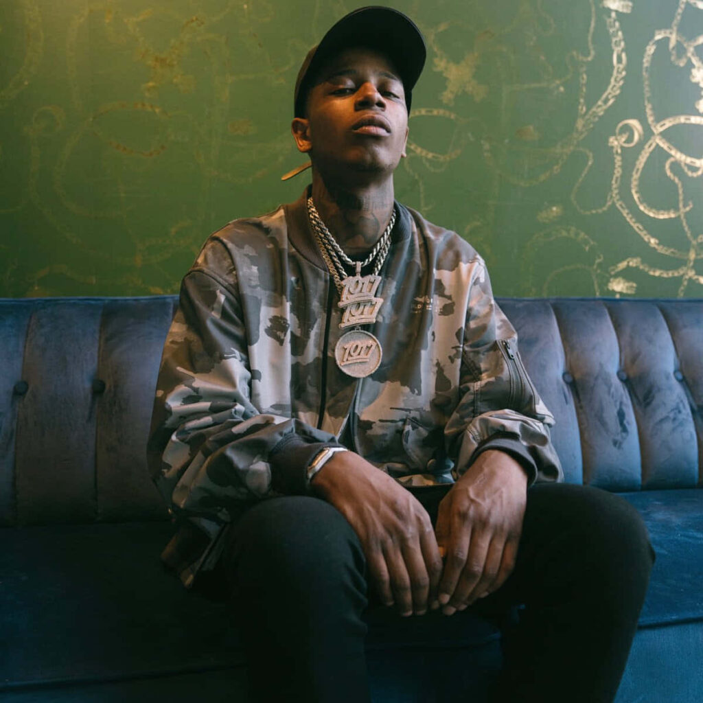 Big Scarr American rapper in camouflage jacket and black cap sitting on blue couch wallpaper background