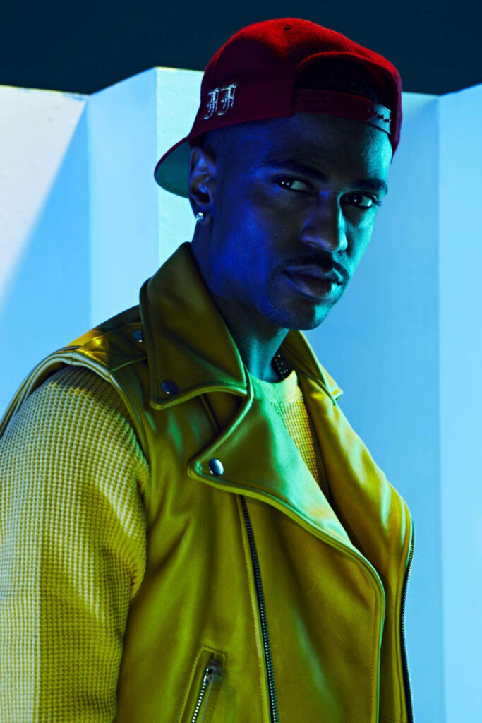Big Sean in bold yellow jacket and red cap against vibrant blue wallpaper background