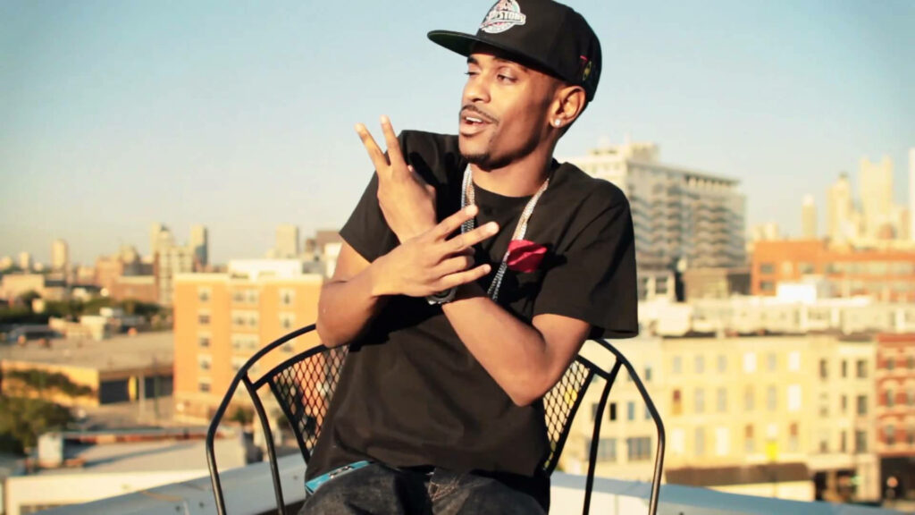 Big Sean sitting outdoors on metal chair showing peace sign wearing black T-shirt and cap urban vibe Wallpaper