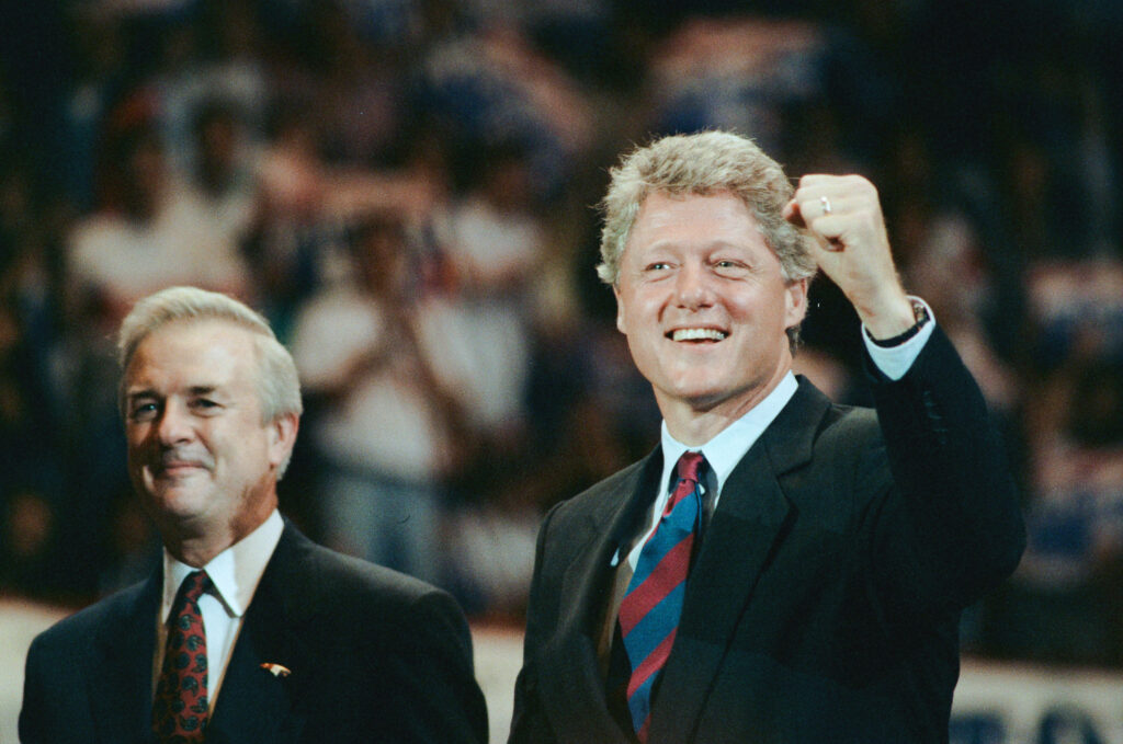Bill Clinton smiling and raising fist in celebration before cheering crowd against wallpaper background