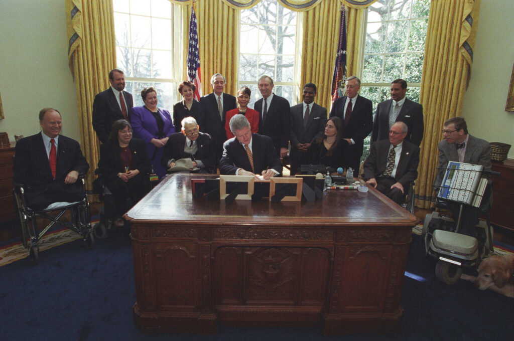 Bill Clinton signing agreement at Resolute desk in Oval Office with diverse supporters united Wallpaper