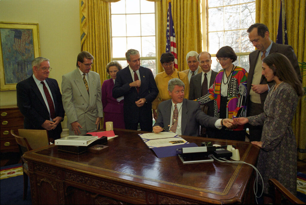 Bill Clinton signing important document in Oval Office surrounded by people historic moment Wallpaper