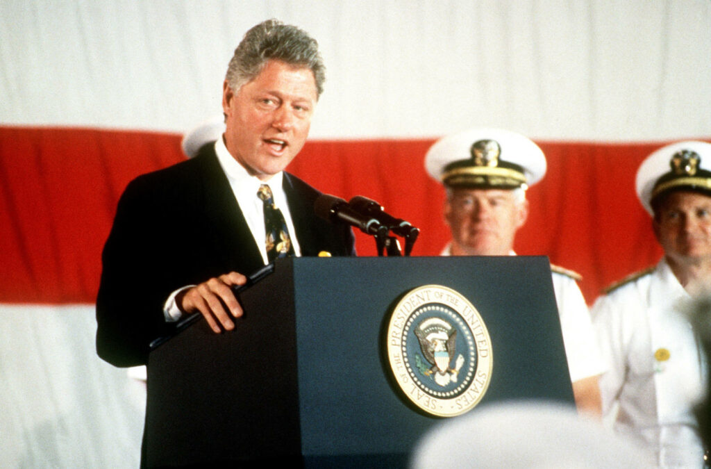 Bill Clinton speaking at podium with military personnel and American flag backdrop wallpaper