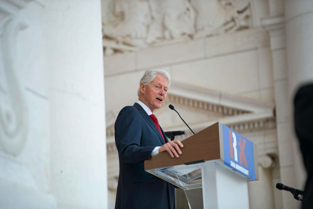 Bill Clinton speaking thoughtfully at podium with elegant architectural wallpaper background