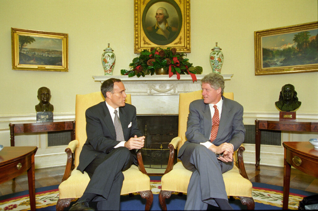 Bill Clinton and Rudy Giuliani talking in Oval Office with historic portraits and elegant decor Wallpaper