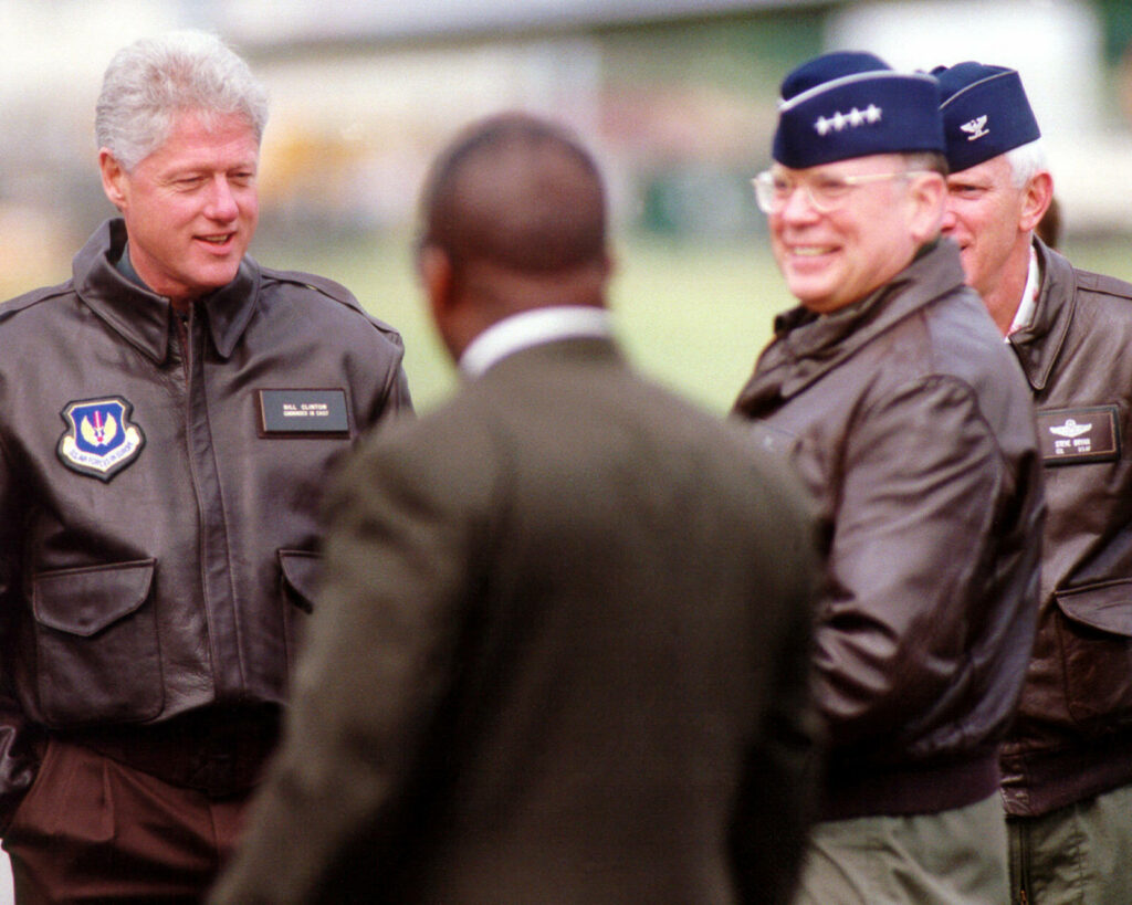 Bill Clinton in flight jacket outdoors talking with military officials dynamic authoritative scene Wallpaper
