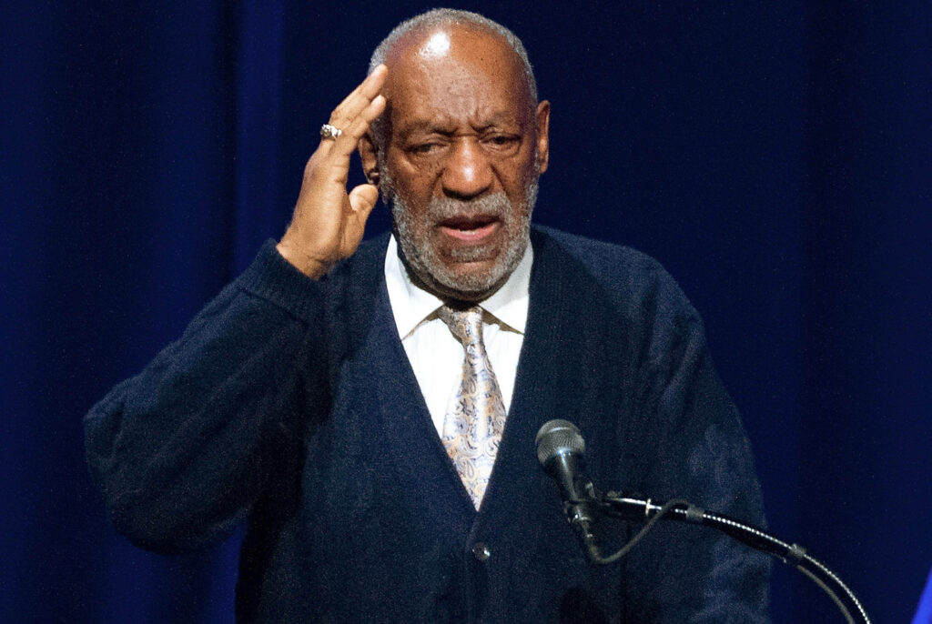 Bill Cosby delivering passionate speech on stage wearing dark sweater and patterned tie Wallpaper