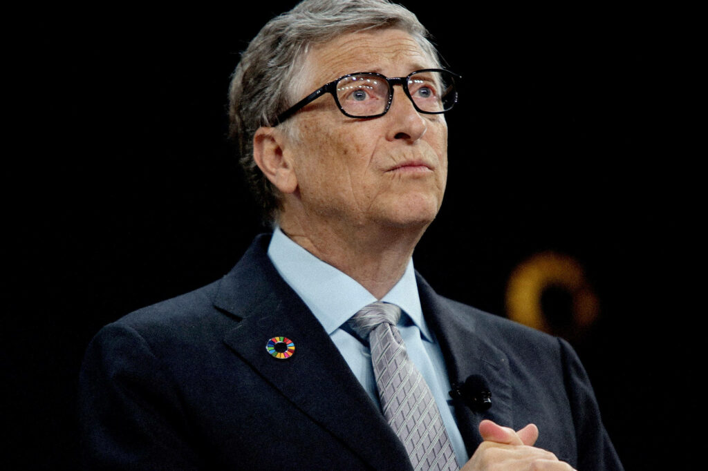 Bill Gates in suit with LGBTQ pin on lapel standing thoughtfully against wallpaper background