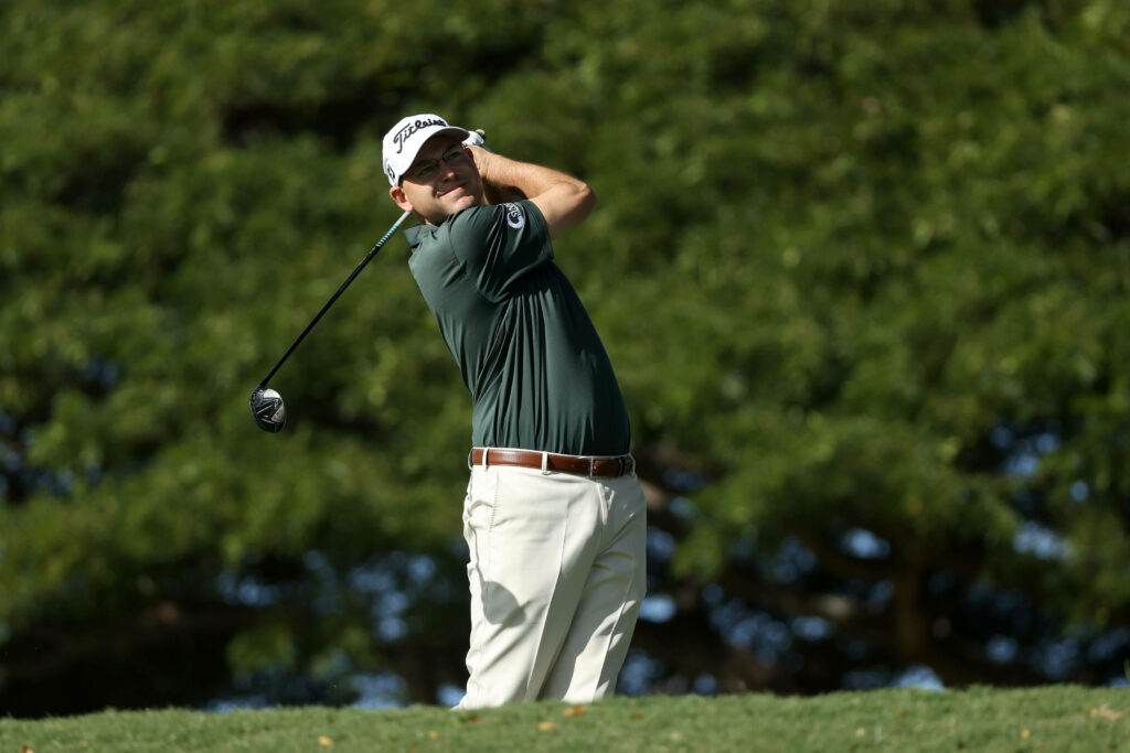Bill Haas mid golf swing in dark green shirt white pants with lush trees golf course background Wallpaper