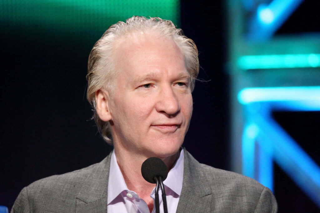 Bill Maher in gray suit standing confident with colorful illuminated background wallpaper