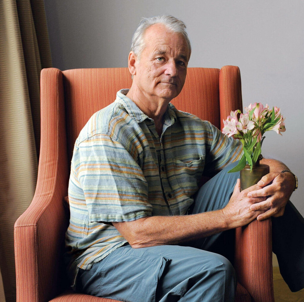 Bill Murray relaxed in orange chair holding flowers in softly lit hotel room with wallpaper background