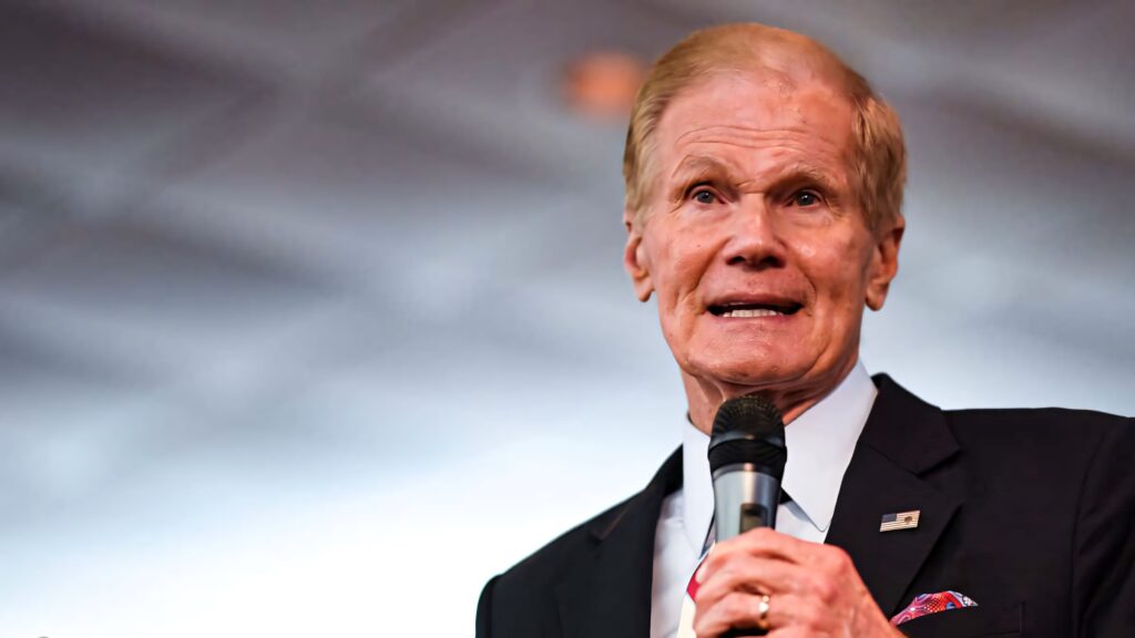 Bill Nelson in suit holding microphone ready to speak gray patterned wallpaper background