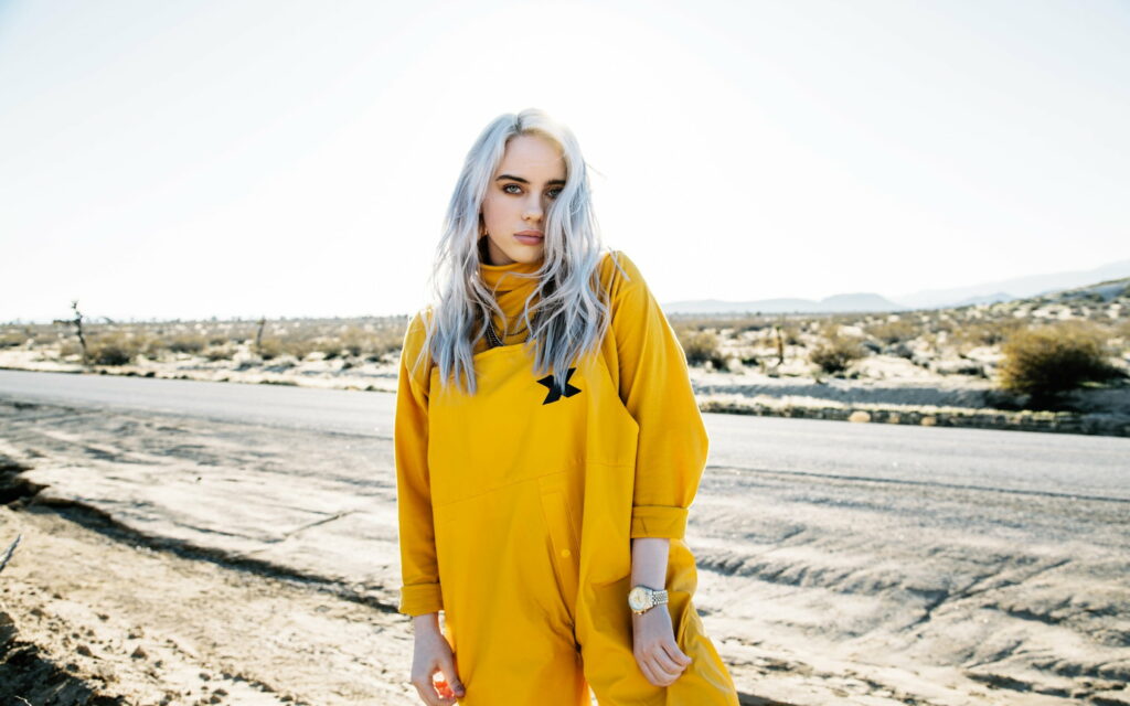 Billie Eilish in vibrant yellow jumpsuit with platinum hair posing in sunlit desert background Wallpaper
