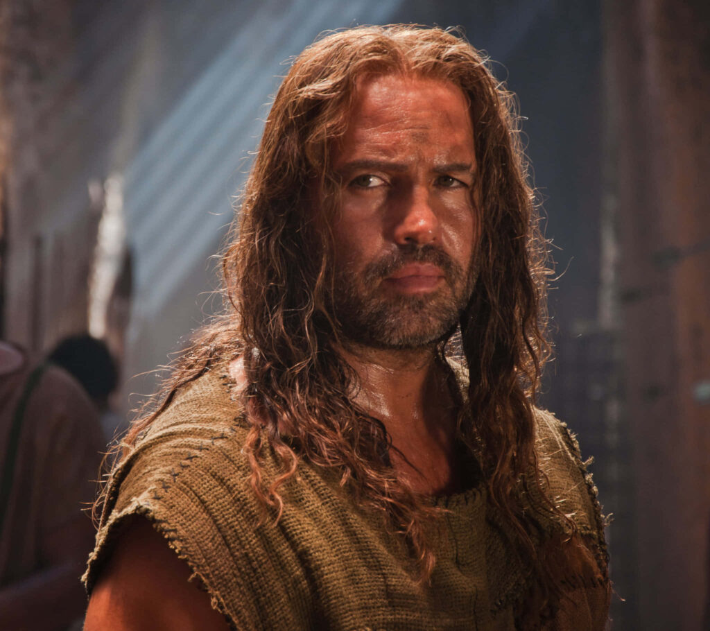 Billy Zane with long hair and rugged attire in dimly lit setting with sunlight rays wallpaper background