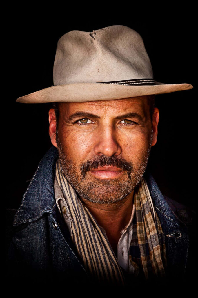 Billy Zane portrait wearing light fedora and patterned scarf on dark dramatic stylish wallpaper background