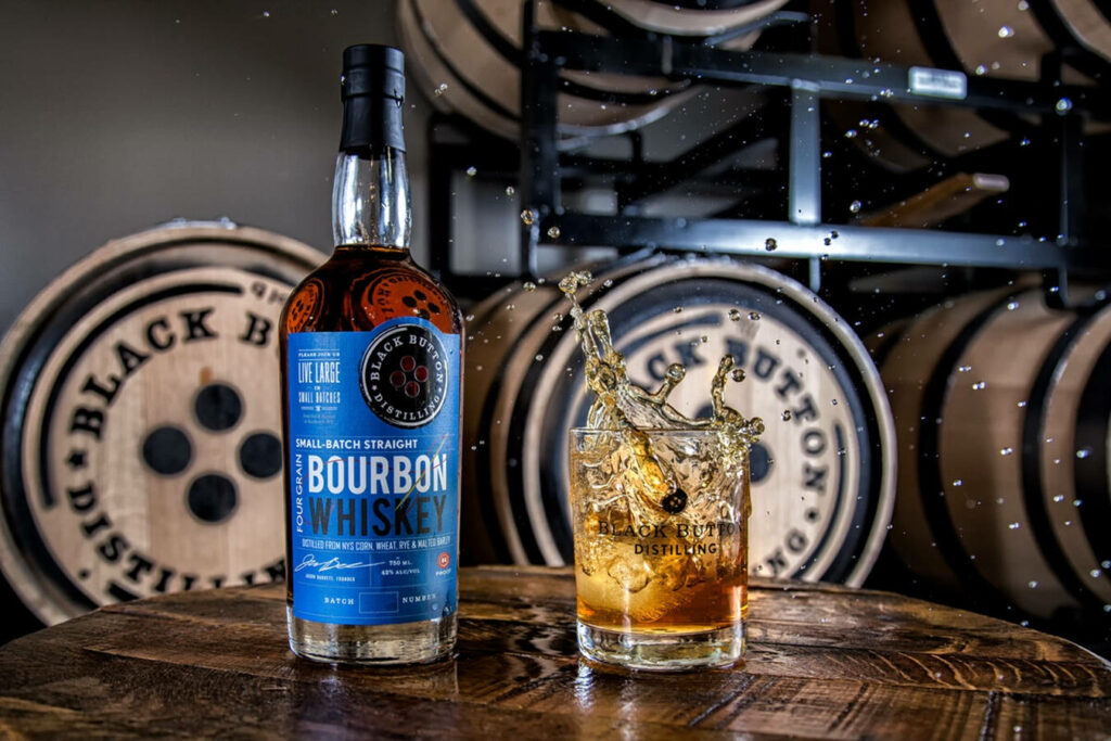 Black Button Distilling bourbon bottle and splash glass with ice in front of branded barrels Wallpaper