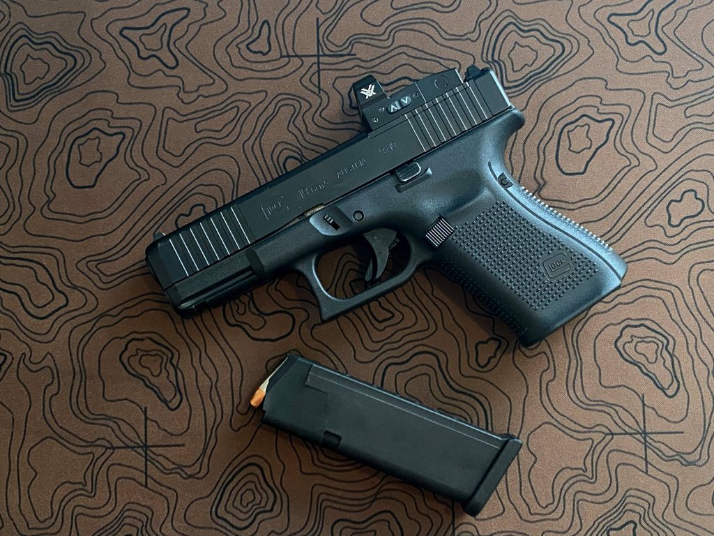 Top-down view of black Glock 19 Gen 5 with Vortex red dot on map mat next to magazine Wallpaper