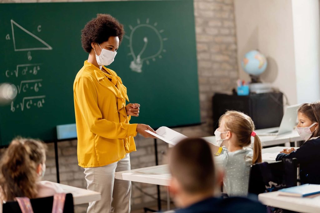 Black teacher in yellow blouse handing worksheet to masked student by math chalkboard classroom Wallpaper