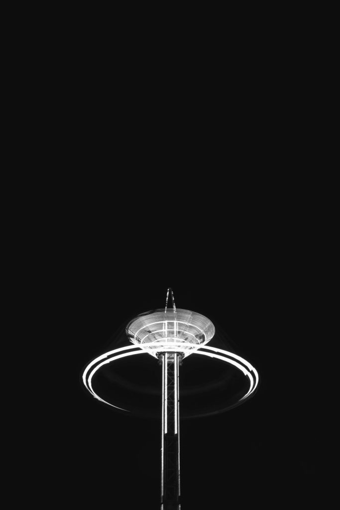Black and white long exposure spinning amusement park tower ride with glowing light trails Wallpaper