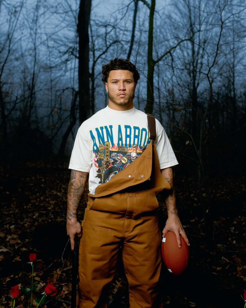 Blake Corum in brown overalls holding football in misty forest showing determination and athleticism Wallpaper