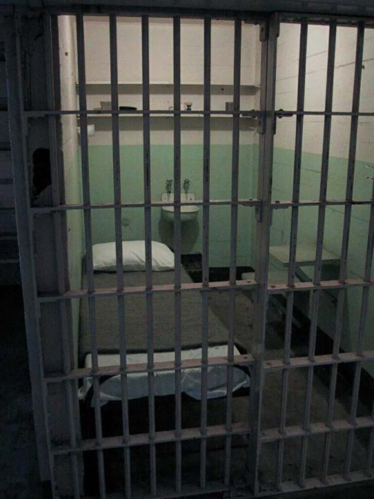 Bleak prison cell with steel bars dim lighting spartan bed and wall-mounted sink wallpaper background
