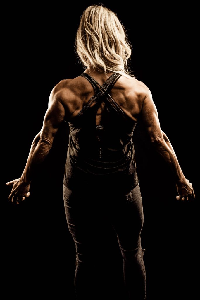 Blonde female bodybuilder back view with dramatic lighting showing muscular definition black background Wallpaper