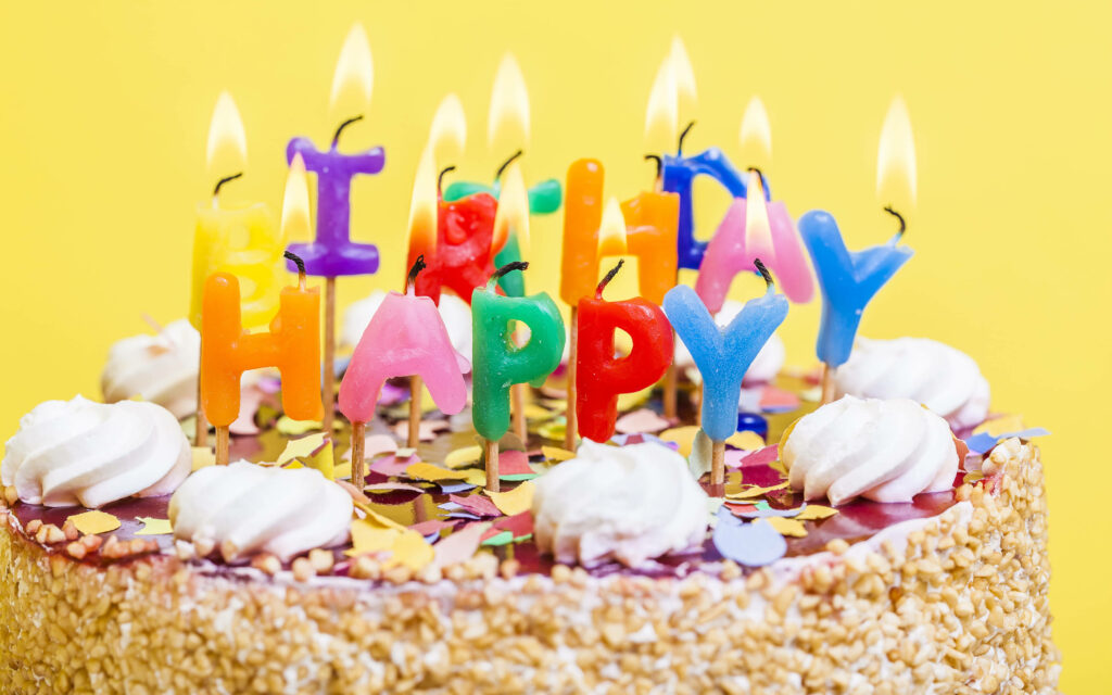 Bright birthday cake with glowing candles spelling HAPPY BIRTHDAY on yellow confetti background Wallpaper