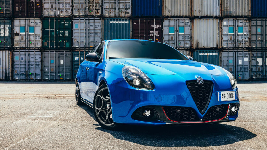 Blue Alfa Romeo Giulietta sportscar parked before colorful stacked shipping containers wallpaper