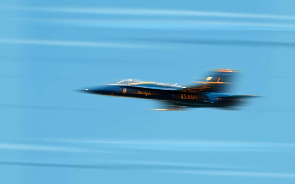 Blue Angels jet soaring fast with motion blur against clear bright blue sky wallpaper background