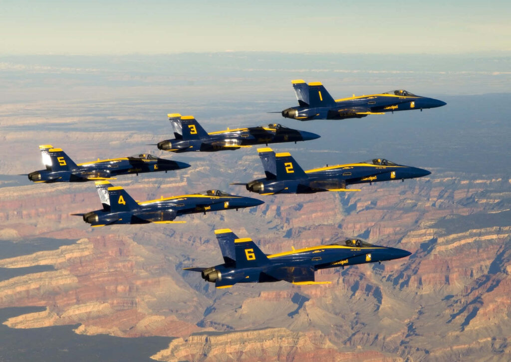 Six Blue Angels jets in blue and yellow fly in tight formation over a scenic canyon background Wallpaper