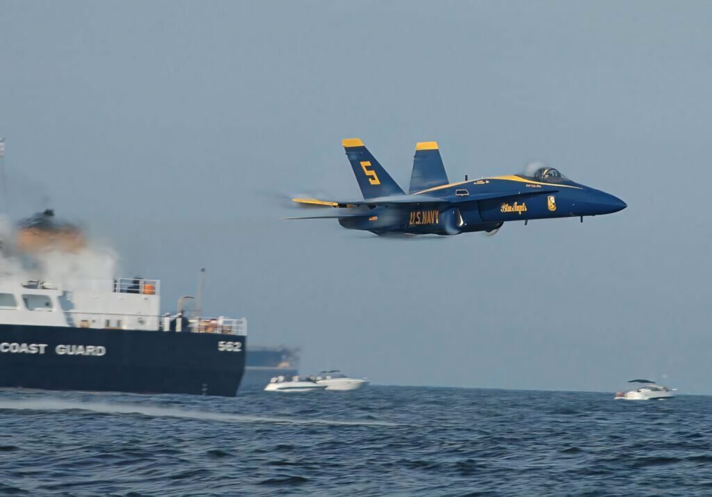 Blue Angels US Navy jet soaring above ocean near Coast Guard ship and boats in flight display Wallpaper