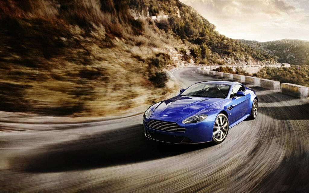 Blue Aston Martin V12 Vantage speeding on winding mountain road high performance sports car wallpaper