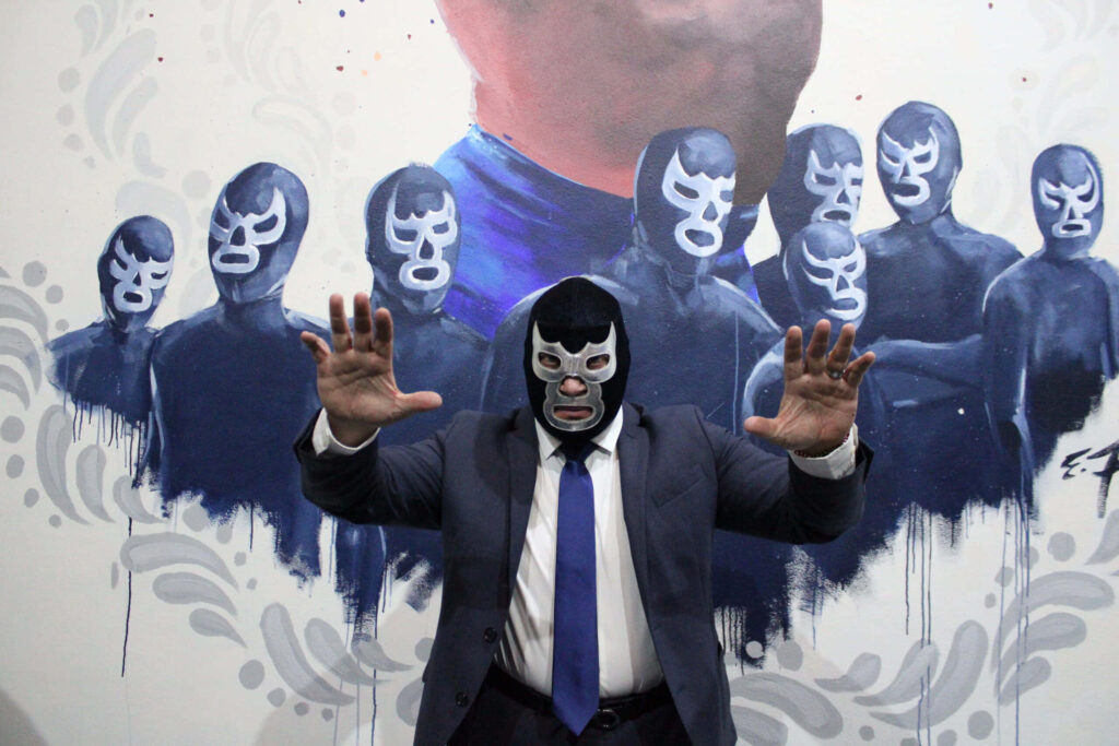 Person in blue demon mask and suit stands before graphic mural with multiple blue masks wallpaper