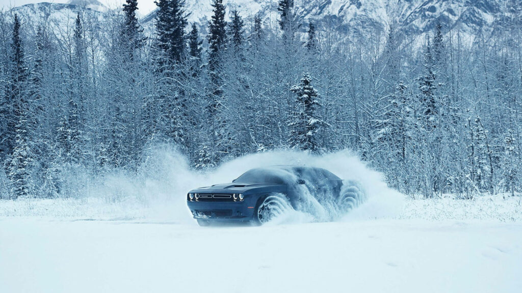 Blue Dodge Challenger speeding through snowy landscape with frosty trees and mountains background Wallpaper