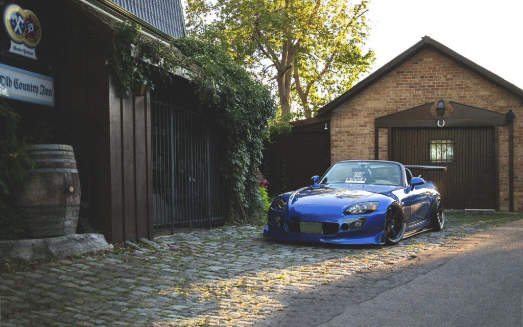 Blue Honda S2000 on old cobblestone street with rustic historical building in sunlit scene wallpaper