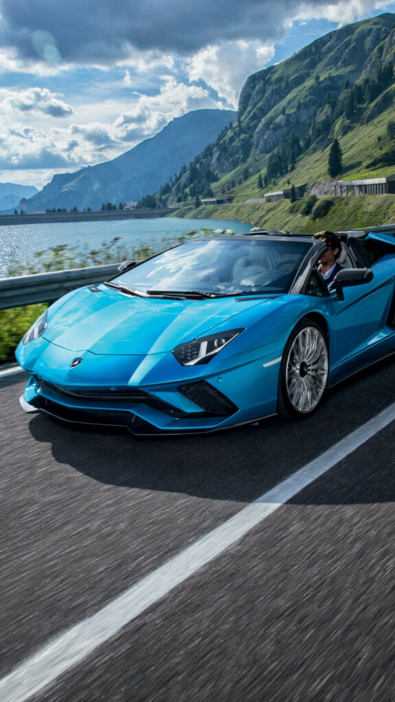 Blue Lamborghini speeding on mountain road beside lake with green hills and dramatic clouds 4k Wallpaper