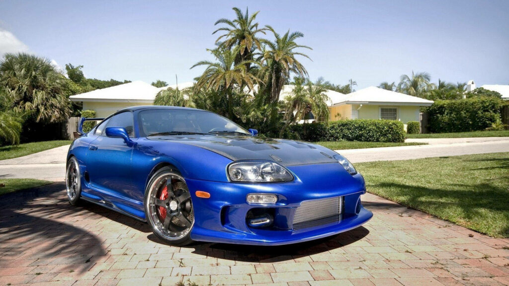 Blue Toyota Supra Mk4 with carbon fiber hood parked on brick driveway near palm trees and houses Wallpaper